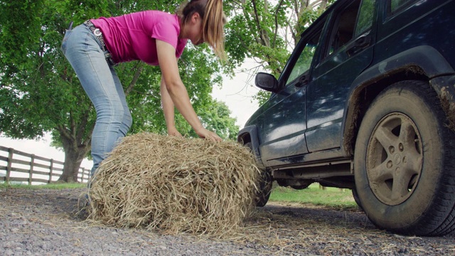一个二十多岁的年轻女人下了车，把一捆干草从屋顶上滚下来，运到农场视频素材