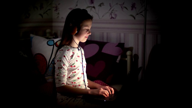 十几岁的女孩，穿着睡衣，戴着耳机，在笔记本电脑上看东西，打字，听音乐，晚上在卧室里。孩子，青少年晚上用笔记本电脑工作视频下载