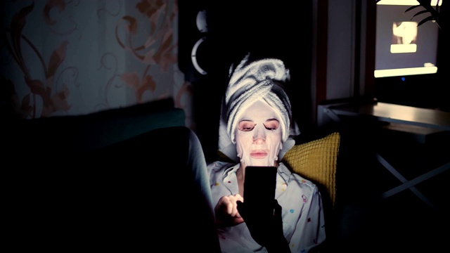 年轻女子戴着化妆布面具，头上戴着白色毛巾，手持智能手机，听着，看着，晚上，躺在沙发上，在手机屏幕前视频下载