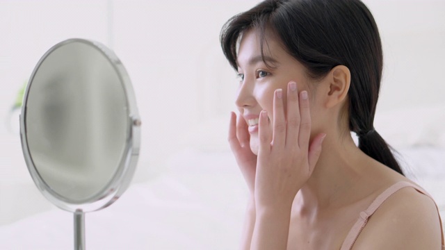 美丽的年轻亚洲女人微笑看着镜子检查脸与皮肤护理和美容年轻化和卫生，美丽的女孩快乐清洁面部与霜或乳液和健康。视频素材