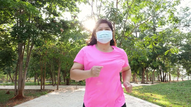 亚洲胖女人美丽的脸晨练在公园跑步戴口罩，防止病毒感染通过呼吸系统，阳光在背景视频下载