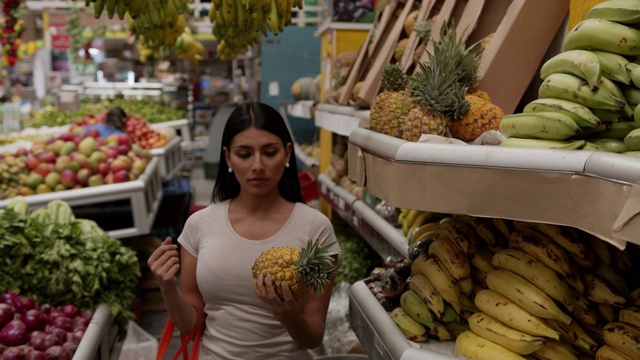 一位漂亮的顾客在市场上拿着可重复使用的袋子从架子上抓菠萝视频素材