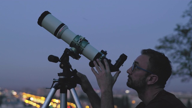 天文学家用望远镜观察星星和以模糊的城市灯光为背景的月亮。视频下载