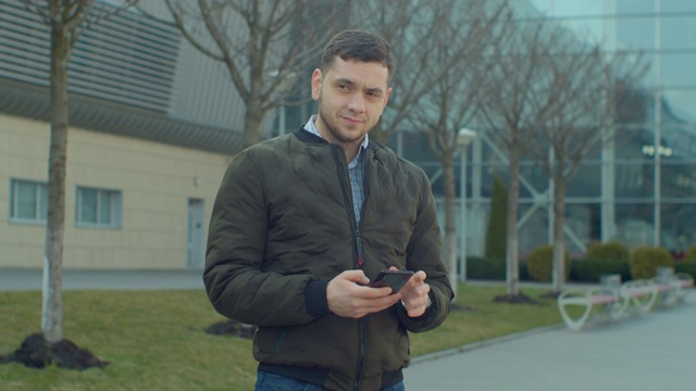 一个男人带着智能手机走在现代办公中心附近。在社交网络上和朋友聊天。看起来很满意，很愉快。拥有现代移动设备。视频素材