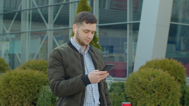一个男人带着智能手机和耳机在现代办公中心附近行走的肖像。在社交网络上和朋友聊天。看起来很满意，很愉快。拥有现代移动设备。视频素材