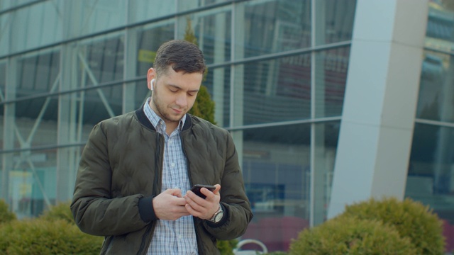 一个男人带着智能手机和耳机在现代办公中心附近行走的肖像。在社交网络上和朋友聊天。看起来很满意，很愉快。拥有现代移动设备。视频素材