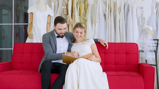 年轻的夫妇休闲看书坐在婚纱店的沙发上为选择最好的婚纱。视频下载