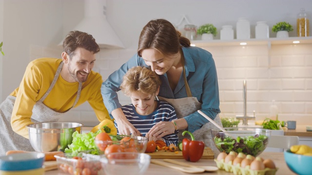 在厨房:三口之家一起烹饪健康晚餐。妈妈和爸爸教小男孩健康的习惯和切蔬菜做晚餐沙拉。可爱的孩子帮助他们美丽的父母视频素材