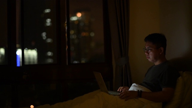 一个亚洲华人中年人晚上在家工作，在床上用他的笔记本电脑视频下载