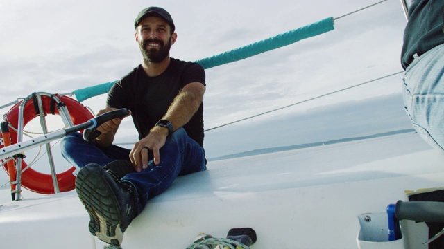 慢动作拍摄的一个白人男子在他的30岁微笑，因为他坐在一艘帆船的甲板上，并在普吉特湾在一个多云的天在华盛顿驾驶它视频素材