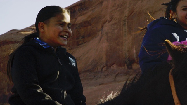 一个10岁的印第安男孩(纳瓦霍人)微笑和大笑，而坐在他的马旁边的一个十几岁的女孩在纪念碑谷沙漠在亚利桑那州/犹他州在一个阳光灿烂的日子旁边的一个大的岩石组视频下载