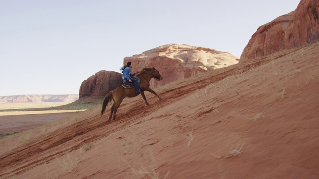 一个十几岁的美国土著女孩(纳瓦霍人)骑着她的马在一个陡峭的，桑迪山在纪念碑谷沙漠在亚利桑那州/犹他州在日落旁边的一个大的岩层视频下载
