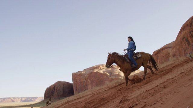 一个十几岁的美国土著女孩(纳瓦霍人)骑着她的马与她的狗在亚利桑那纪念碑谷沙漠在日落旁边的一个大的岩石形成陡峭的小山视频下载