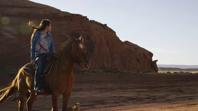 一个十几岁的美国土著女孩(纳瓦霍人)坐在她的马和看风景的纪念碑谷沙漠在亚利桑那州/犹他州在日落旁边的一个大的岩层视频下载