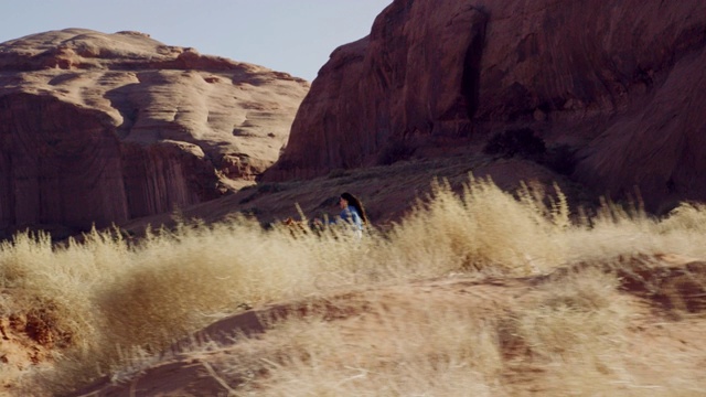 在一个阳光明媚的日子里，10岁的美国土著男孩(纳瓦霍人)骑着马在亚利桑那州的纪念碑谷沙漠追赶一个十几岁的女孩，她和她的宠物狗在一个大的岩层旁边视频下载