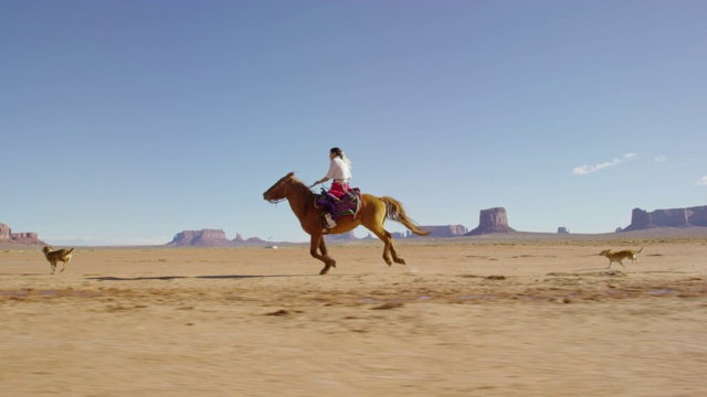 在一个晴朗晴朗的日子里，一个十几岁的印第安女孩穿着传统的纳瓦霍服装骑着她的马疾驰在纪念碑谷沙漠和她的宠物狗视频下载