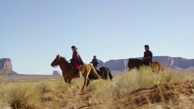 几个年轻的印第安人(纳瓦霍)儿童骑着马通过纪念碑谷沙漠与他们的宠物狗在亚利桑那州/犹他州在一个晴朗，明亮的日子视频下载