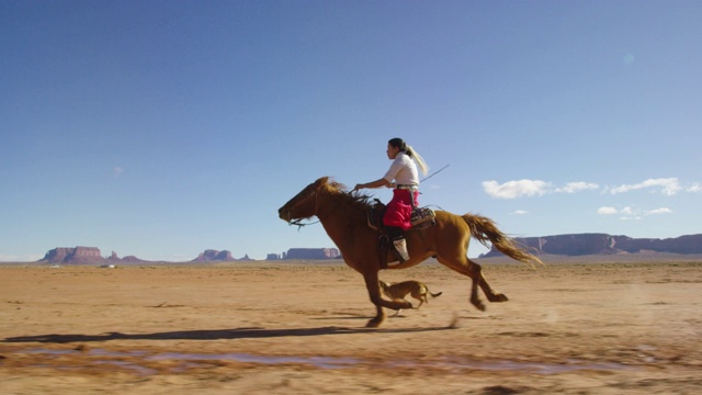 在一个晴朗晴朗的日子里，一个十几岁的印第安女孩穿着传统的纳瓦霍服装骑着她的马疾驰在纪念碑谷沙漠和她的宠物狗视频下载