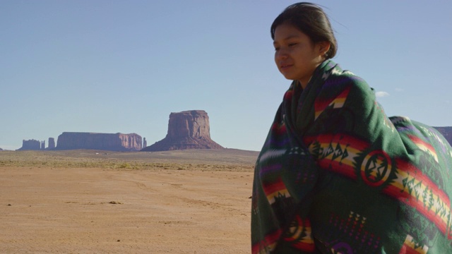 一个十几岁的美国土著女孩在一个晴朗、明亮的日子里，在纪念碑谷沙漠与巨大的岩层之间，用传统的纳瓦霍毯子裹着她的肩膀的慢动作拍摄视频下载