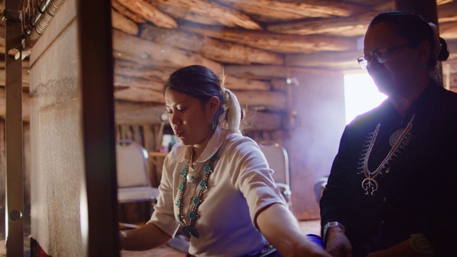 一个美国土著少女在织布机上织布的慢动作镜头，她60多岁的纳瓦霍奶奶在霍根(纳瓦霍木屋)里看着她，并指导她进屋。视频下载