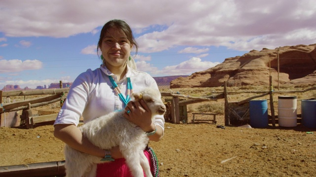 慢动作拍摄的一个十几岁的美国土著女孩抱着一只羔羊和微笑在一个围栏的牧场在纪念碑谷，亚利桑那州/犹他州在一个阳光灿烂的日子与一个大的岩石形成的背景视频下载