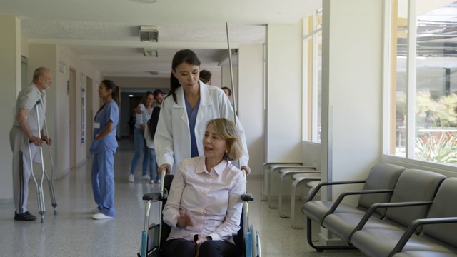 美丽的女医生推着轮椅和女老年病人，在医院的走廊上愉快地交谈视频素材
