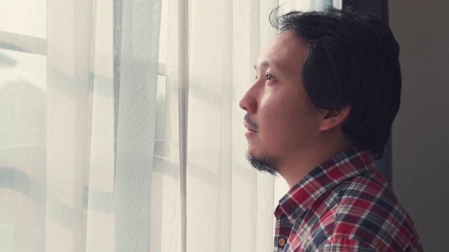 一名亚洲男子正在通过窗口进行自我隔离以检测冠状病毒视频下载