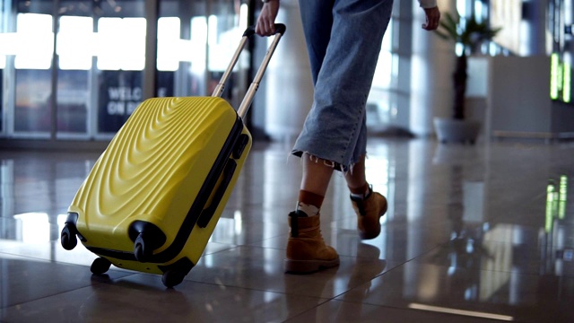 时尚的女人走着小车黄色的箱子由空空如也的机场，低半的女性纤细的腿和中等轮式的包，平滑的跟踪拍摄。穿着牛仔裤和棕色靴子。罕见的视图视频素材