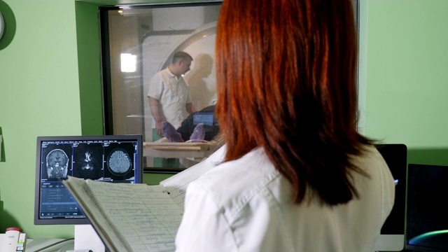 身着白大褂的女性放射科专家看着病人接受CT MRI检查，并在检查x光时写下医疗结果，珀森被磁共振成像扫描仪扫描。高清视频下载