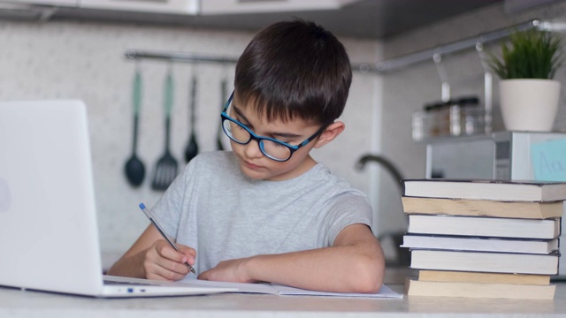 肖像。一个孩子坐在家里厨房的桌子旁用笔记本电脑做作业，用钢笔在笔记本上写字。在线技术视频下载
