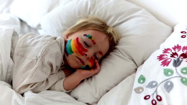 漂亮的金发小男孩，脸上画着彩虹，双手凌乱，睡在床上视频下载