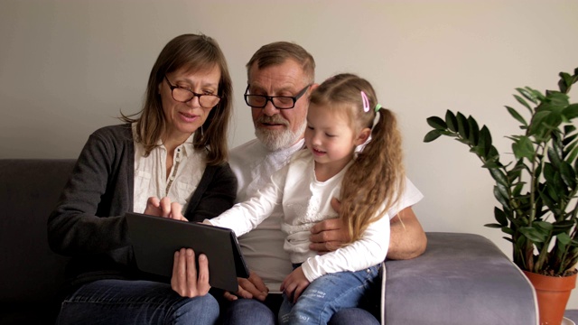 孩子和祖父母在使用平板电脑。一对可爱的中年夫妇和他们的小孙女在家里一起使用数码平板电脑。人才和现代技术视频素材