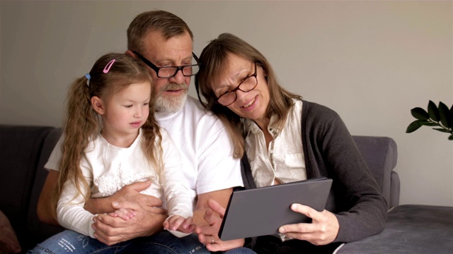 家庭、一代、技术和人的概念——微笑的祖父、孙女和祖母坐在家里的沙发上，拿着平板电脑视频素材