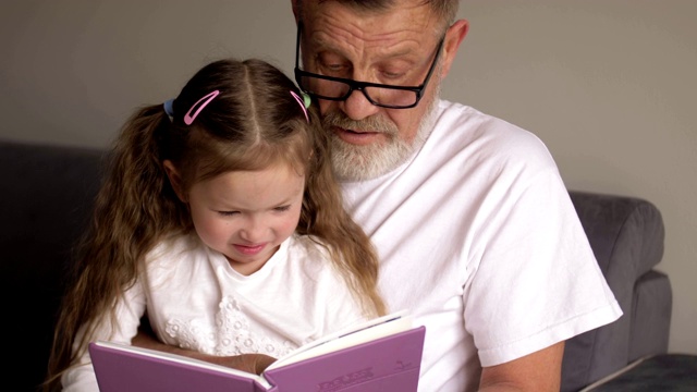 戴眼镜的爷爷和可爱的十几岁的孙女坐在家里的沙发上一起看书视频素材