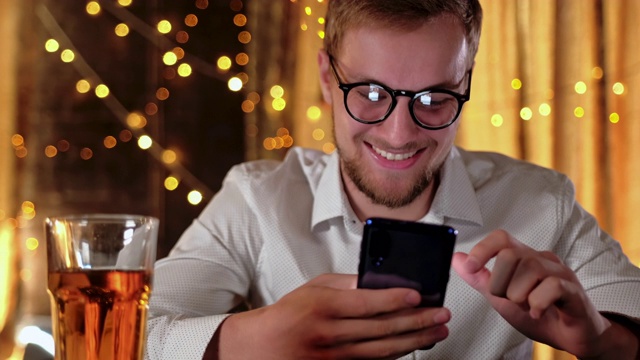 帅气的胡子男子一边在酒吧里喝啤酒一边用智能手机微笑视频素材