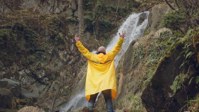 穿着黄色雨衣的年轻男性旅行者正在欣赏美丽的瀑布。在山里远足。徒步旅行者跑向瀑布，举起双手。喜欢的生活方式。概念:宗教、生物、生态。视频素材