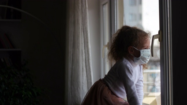 2019冠状病毒病概念，小女孩戴着口罩透过窗户往外看，带着纸条隔离在家。可怜的孩子因为冠状病毒被隔离。视频素材
