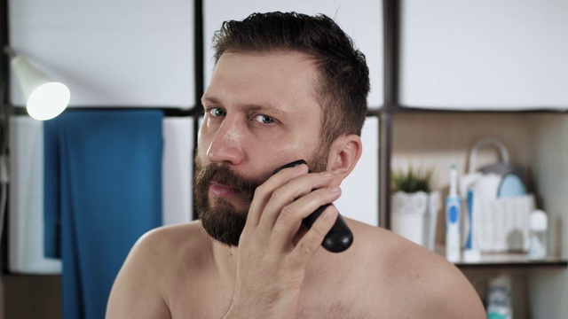 男人把胡子上的头发拉直。有魅力的男人在浴室里看着镜子，用修剪器修剪他的头发，甚至使胡子边缘。美,新鲜的概念。特写镜头视频下载