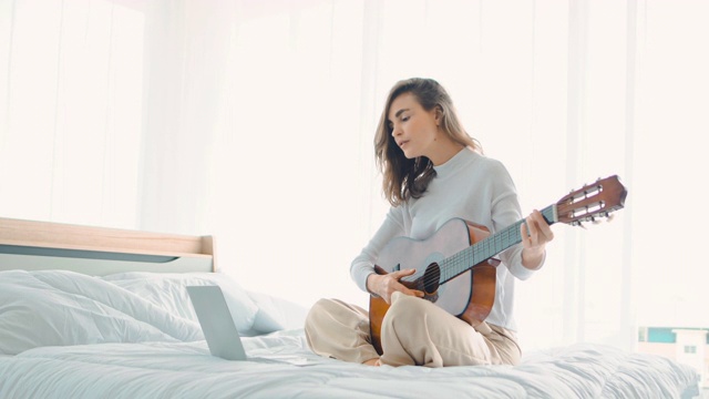 年轻女子集中学习弹吉他使用笔记本电脑坐在家里的床上视频素材