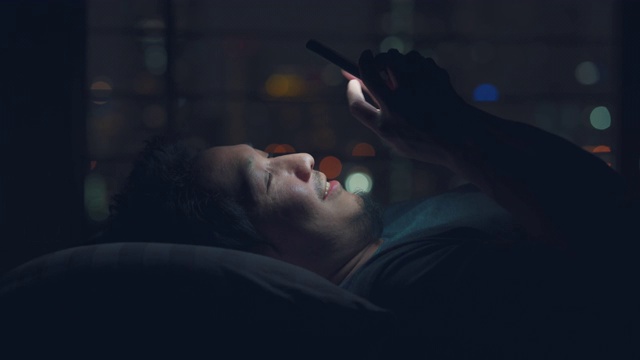 亚洲男人在床上使用智能手机在晚上与散景灯光城市视频素材
