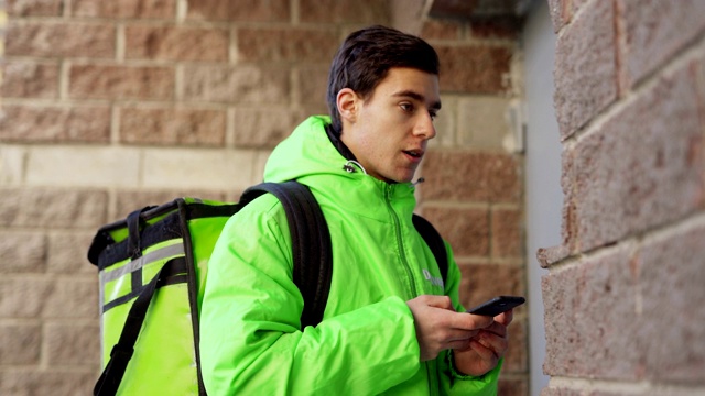 侧视图的年轻男性食品快递在绿色制服与绝缘袋阅读手机上的订单，同时拨打门铃和与客户交谈视频下载