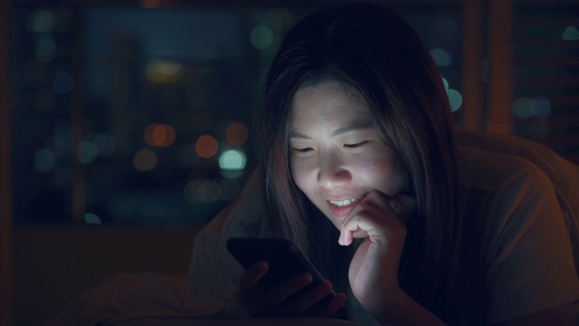 亚洲妇女使用智能手机在床上晚上与散景灯光城市视频素材