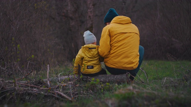 慢镜头:后视镜，父亲坐在一根圆木上给儿子讲故事。快乐的父子坐在公园里聊天。《自然秋景》中穿着黄夹克的父子。视频素材