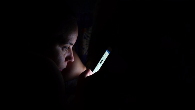 成年女性在黑夜中使用智能手机。年轻女子在家中卧室的床上使用智能手机视频下载