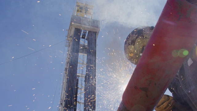 在一个阳光明媚的日子里，一名油田工人在一个石油和天然气钻井平台上的井架旁焊接两根管道，火花四溅视频素材