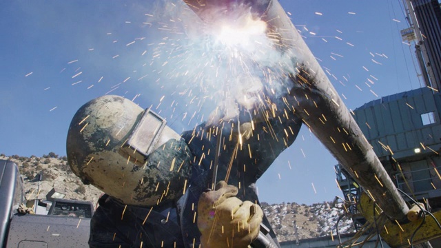 在一个阳光明媚的日子里，一名油田工人在一个石油和天然气钻井平台上的井架旁焊接两根管道，火花四溅视频素材