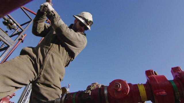 慢镜头拍摄的一个油田工人在他的30多岁泵下管线和敲击管道与一个石油和天然气钻井平台在一个寒冷，阳光，冬天的早晨视频下载