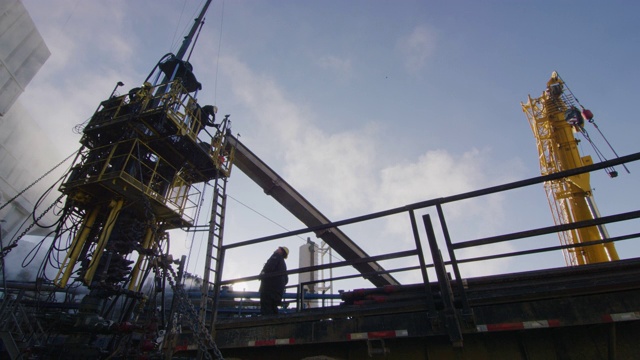 在一个寒冷的冬天的早晨，油田工人在石油和天然气钻井平台的一个高高的金属平台上架设钻杆的慢镜头视频素材