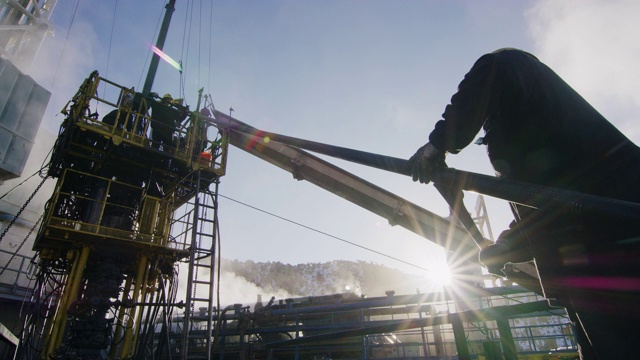 一个寒冷、阳光明媚的冬天早晨，油田工人在石油和天然气钻井平台的一个高高的金属平台上架设钻杆的慢镜头视频素材