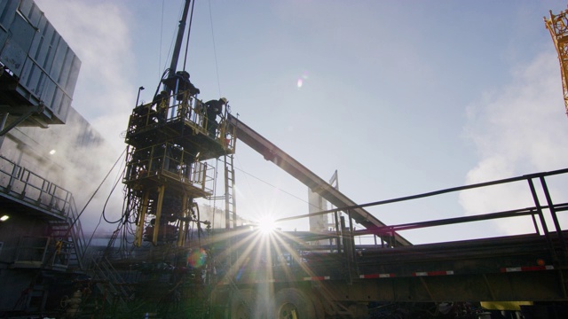 一个寒冷、阳光明媚的冬天早晨，油田工人在石油和天然气钻井平台的一个高高的金属平台上架设钻杆的慢镜头视频素材